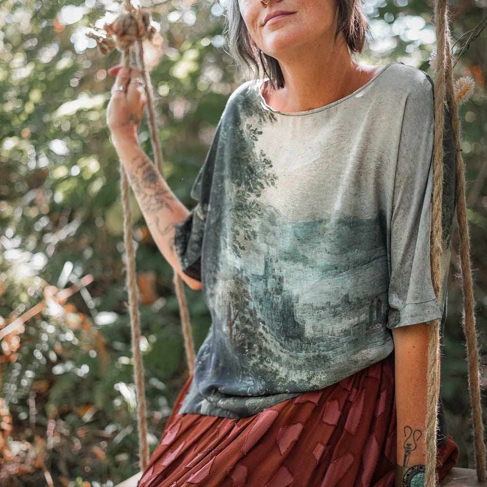 
                  
                    Castles in the Air Luxe Tee featuring Jan Brueghel’s whimsical 1600s landscape and painterly floral trim on soft bamboo-based viscose – Market of Stars.
                  
                