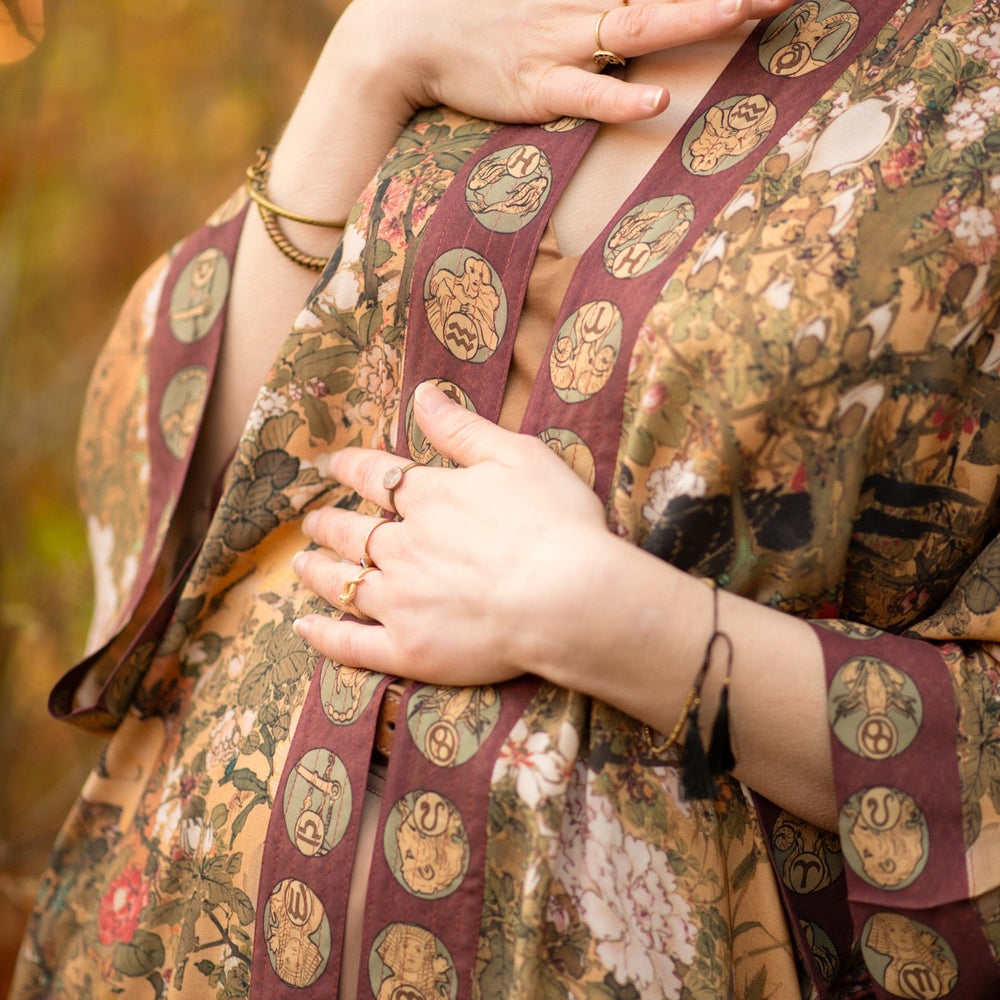 
                  
                    Model wearing Dreamweaver Opera Duster – featuring Alphonse Mucha’s zodiac woman, celestial crown, and burgundy zodiac trim inspired by 1970s luxe bohemian style – Market of Stars.
                  
                