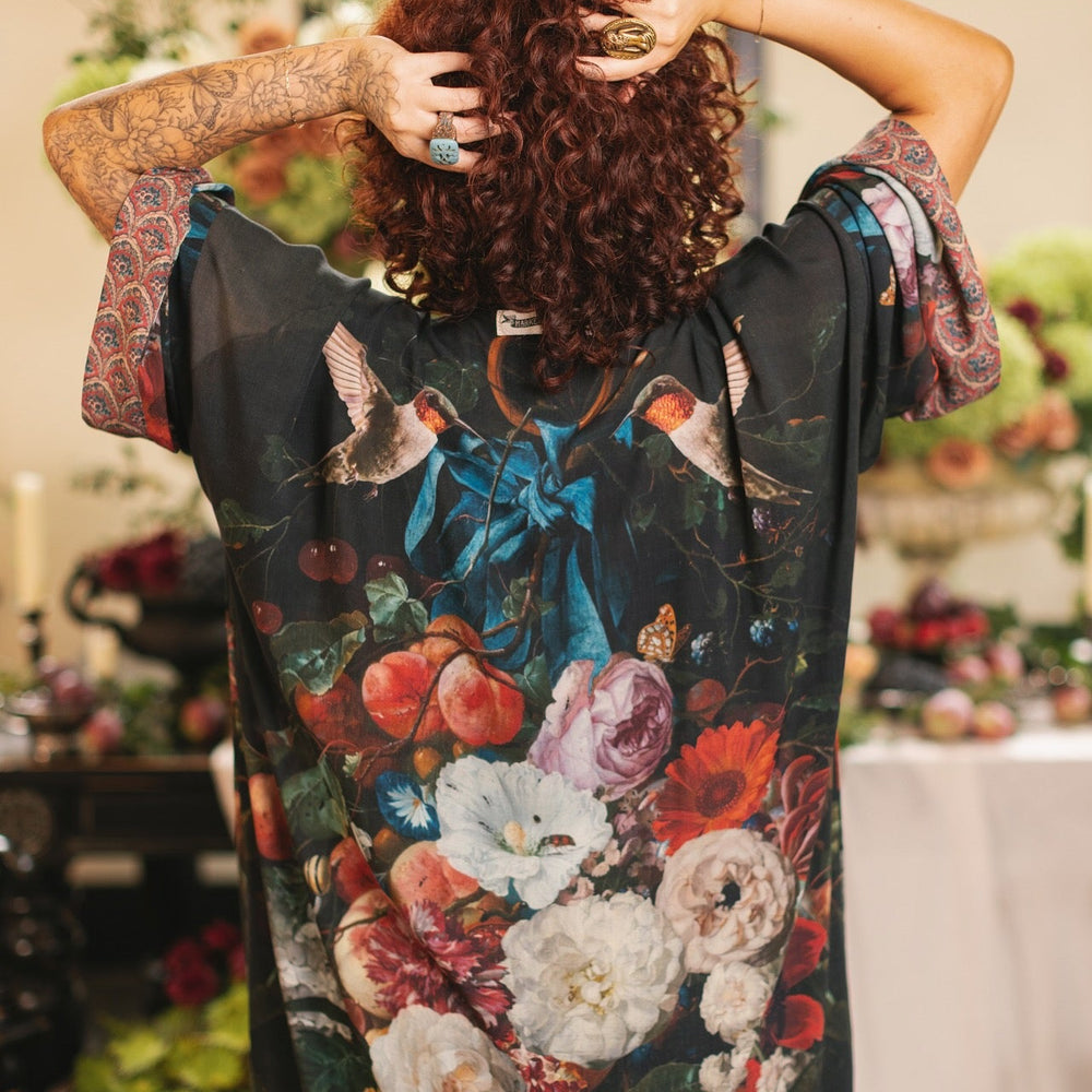 
                  
                    Model showcasing the Flight of Fancy Opera Robe featuring vibrant hummingbirds and historic floral paintings by Rachel Ruysch – Market of Stars.
                  
                