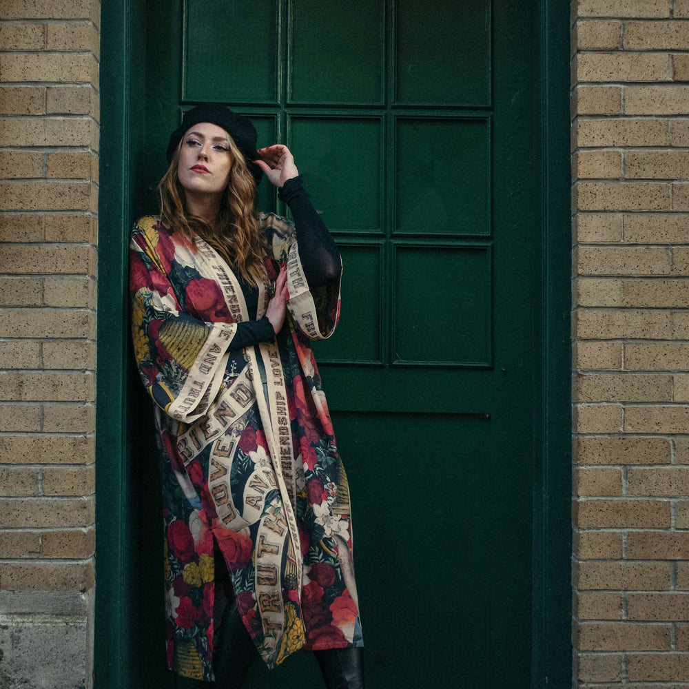 
                  
                    Model wearing our long, eclectic opera duster featuring vintage symbols of love, kindness, and truth in a bold archive-inspired print with removable belt – Market of Stars.
                  
                