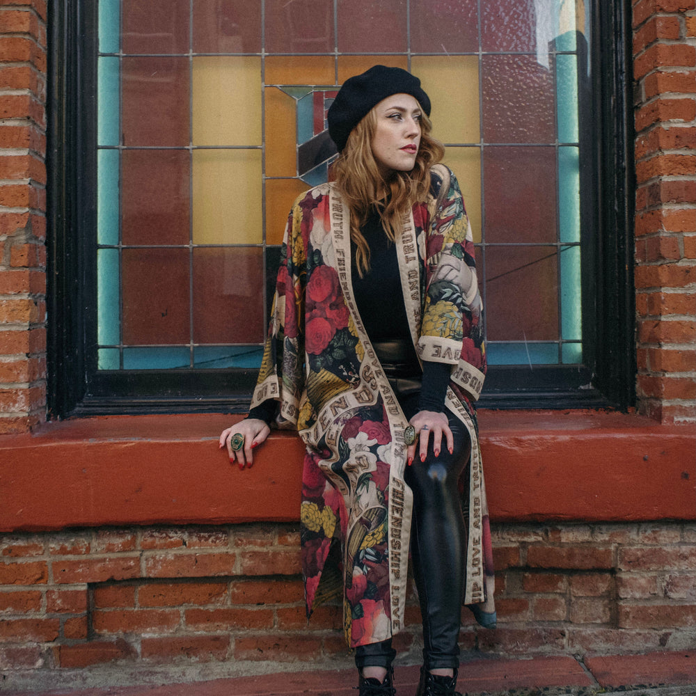 
                  
                    Model wearing our long, eclectic opera duster featuring vintage symbols of love, kindness, and truth in a bold archive-inspired print with removable belt – Market of Stars.
                  
                