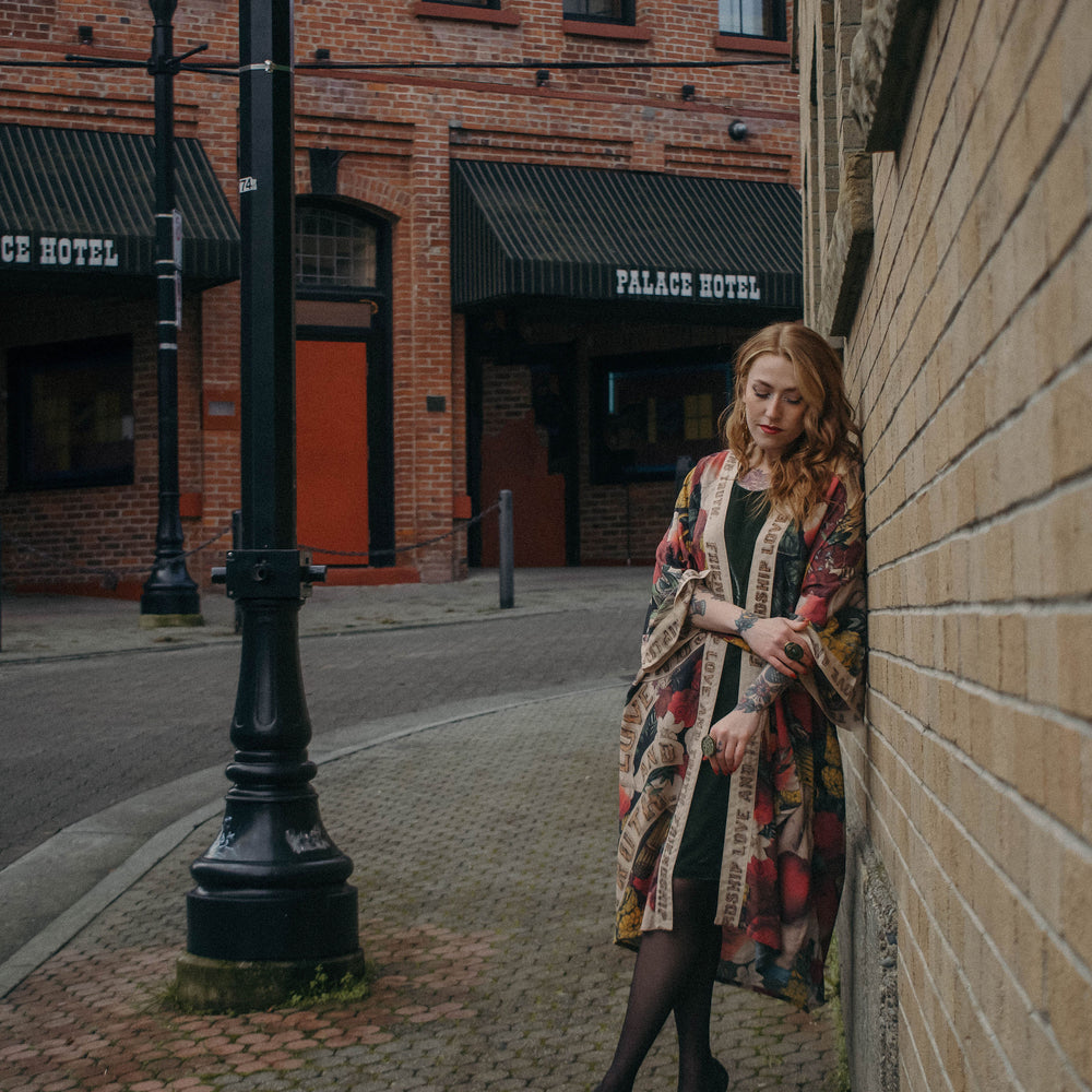 
                  
                    Model wearing our long, eclectic opera duster featuring vintage symbols of love, kindness, and truth in a bold archive-inspired print with removable belt – Market of Stars.
                  
                