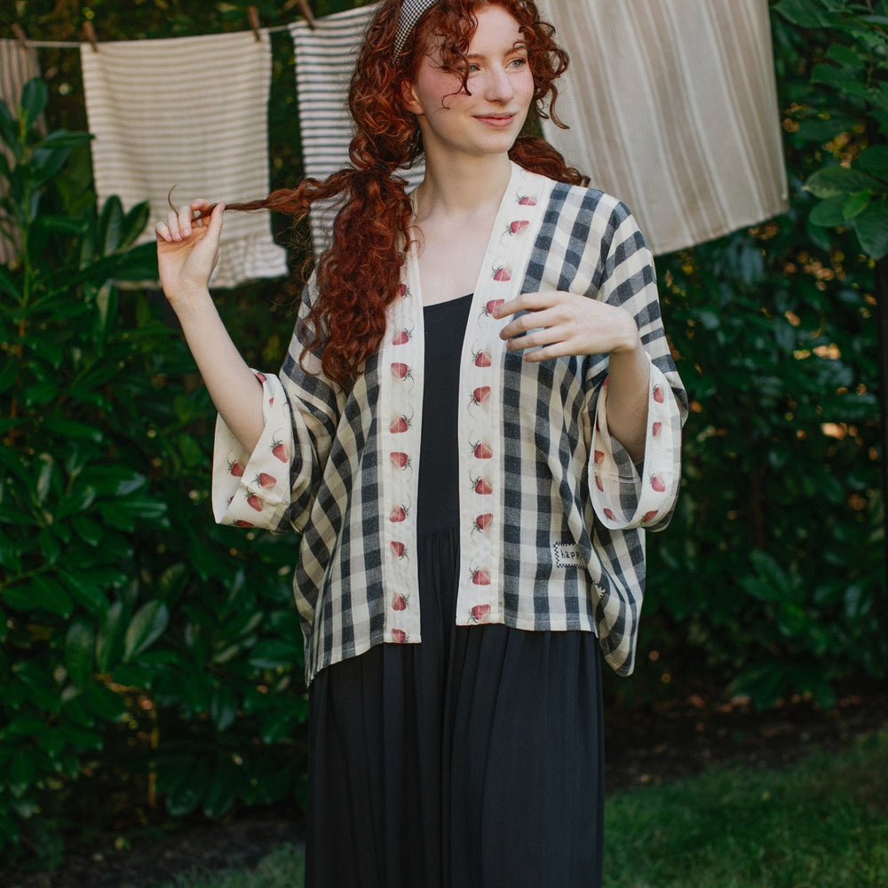 
                  
                    Model wearing Happy Days pixie cut kimono featuring heritage gingham, vintage strawberries, and a cross-stitch patch inspired by an 1836 sampler – Market of Stars.
                  
                