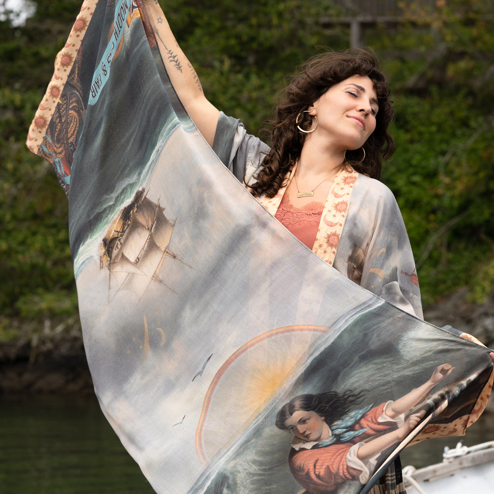 Model wearing the Hold Fast bamboo viscose scarf featuring a Victorian woman bravely rowing through a storm, seagulls, and rainbow horizon – Market of Stars.