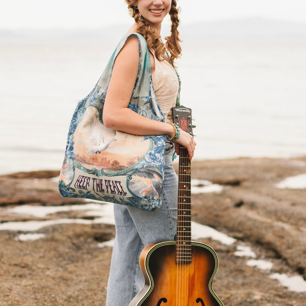 Model wearing our Peace Out tote bag featuring 1800's perfumeur sign with gentle tones of blue & aqua, landscapes, clouds, dove, and peace symbols -Market of Stars.
