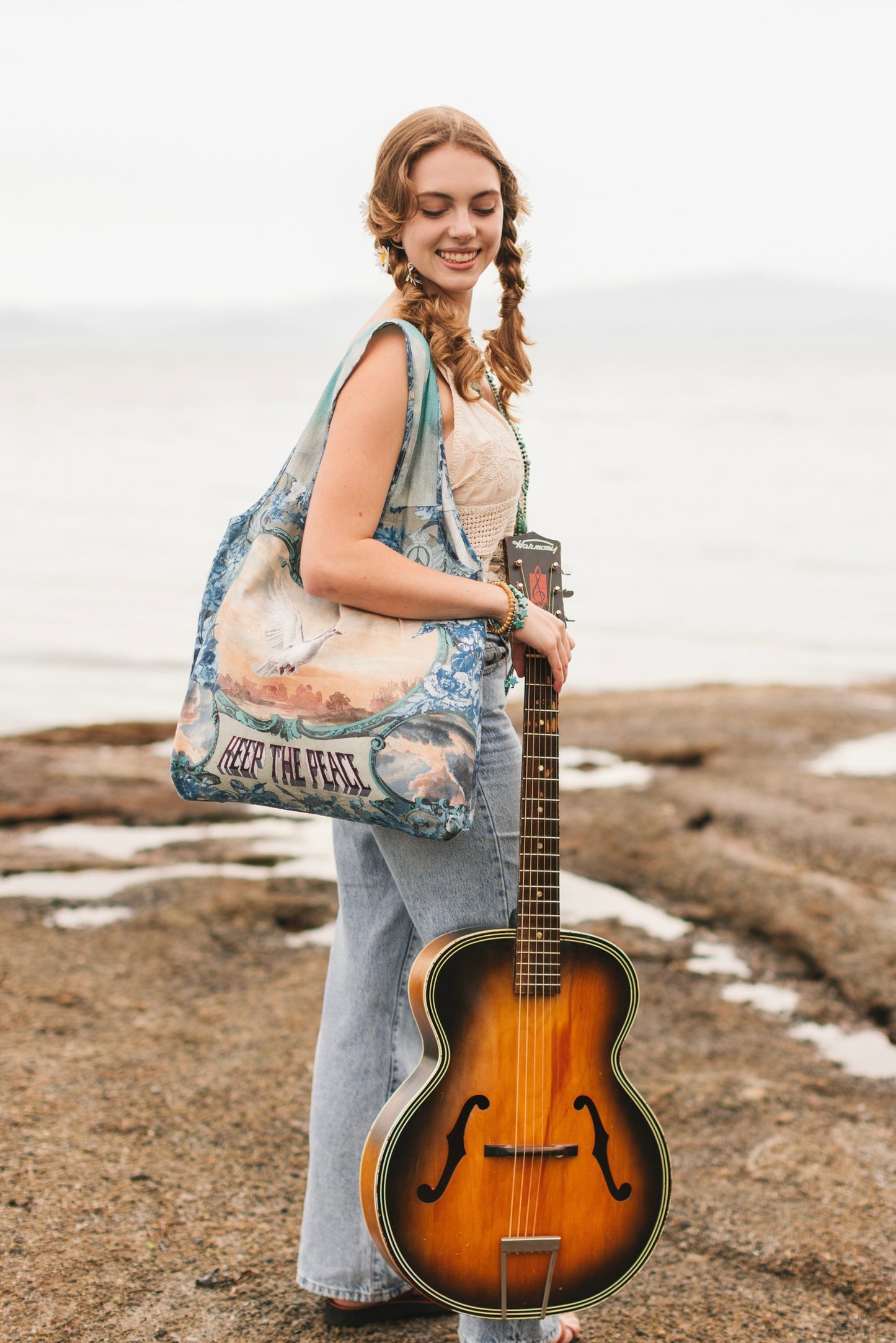Model wearing our Peace Out tote bag featuring 1800's perfumeur sign with gentle tones of blue & aqua, landscapes, clouds, dove, and peace symbols -Market of Stars.