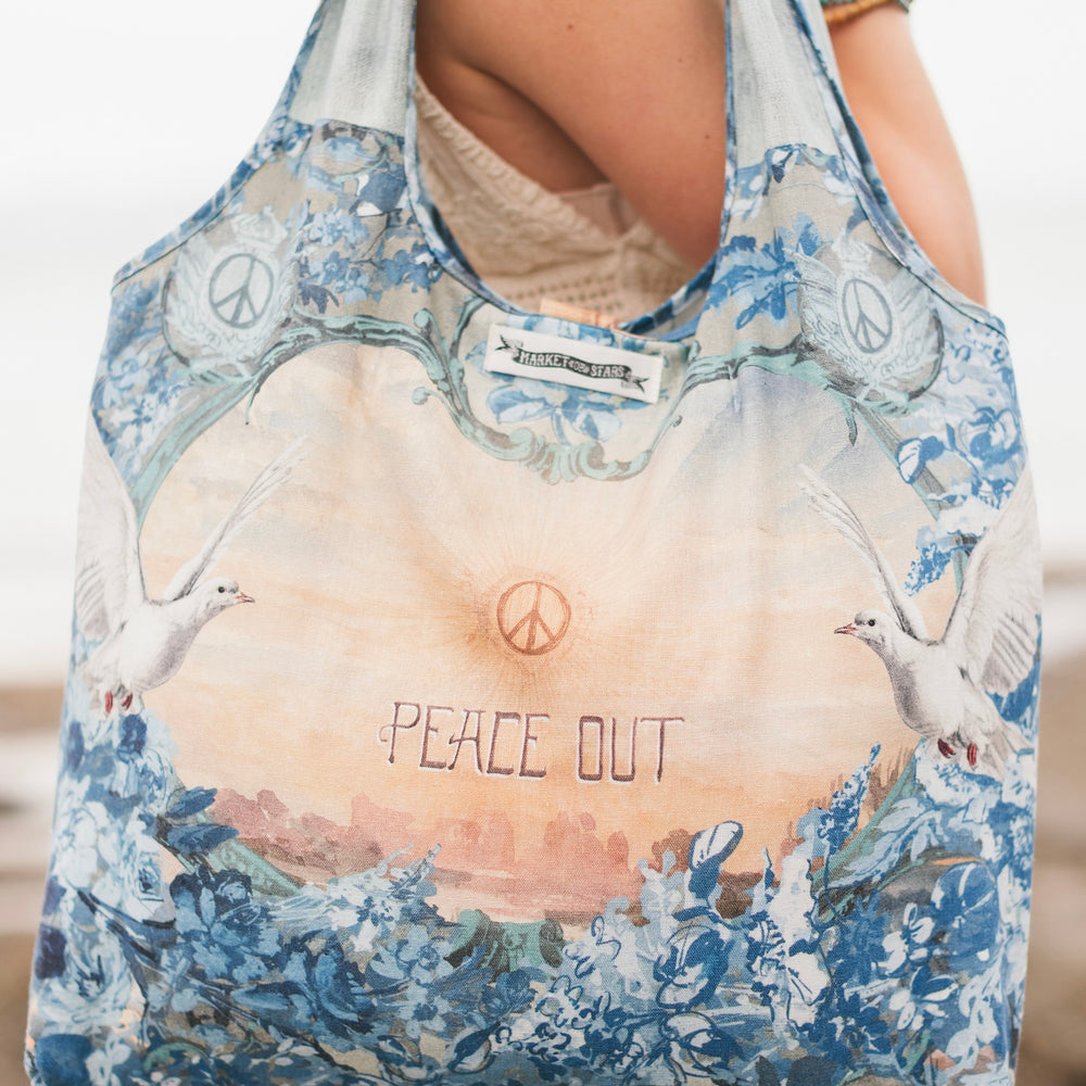 
                  
                    Model wearing our Peace Out tote bag featuring 1800's perfumeur sign with gentle tones of blue & aqua, landscapes, clouds, dove, and peace symbols -Market of Stars.
                  
                