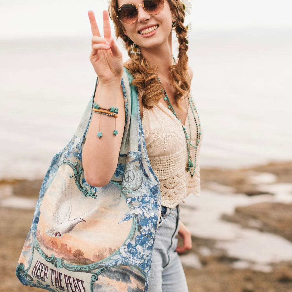 
                  
                    Model wearing our Peace Out tote bag featuring 1800's perfumeur sign with gentle tones of blue & aqua, landscapes, clouds, dove, and peace symbols -Market of Stars.
                  
                