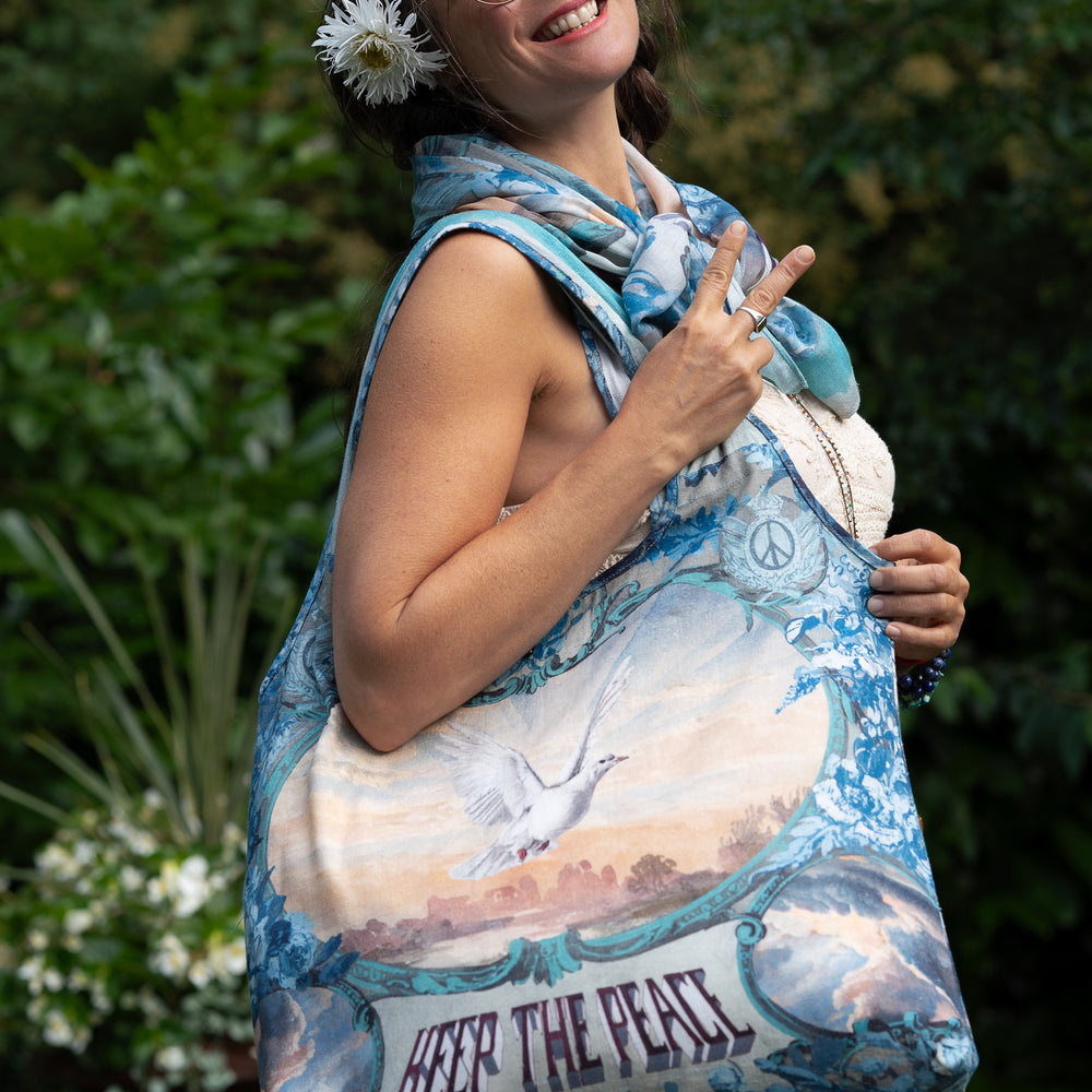 Model wearing our Peace Out tote bag featuring 1800's perfumeur sign with gentle tones of blue & aqua, landscapes, clouds, dove, and peace symbols -Market of Stars.