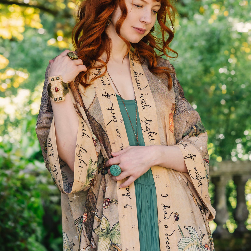 
                  
                    Model wearing an enchanting kimono duster robe featuring illustrations by Charles van Sandwyck's, crafted from airy bamboo-based viscose with a touch of whimsical magic - Market of Stars
                  
                