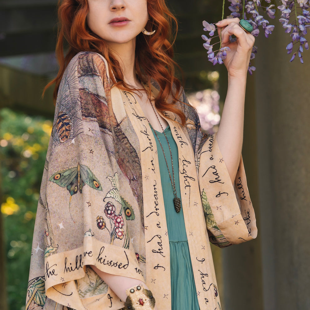 
                  
                    Model wearing an enchanting kimono duster robe featuring illustrations by Charles van Sandwyck's, crafted from airy bamboo-based viscose with a touch of whimsical magic - Market of Stars
                  
                