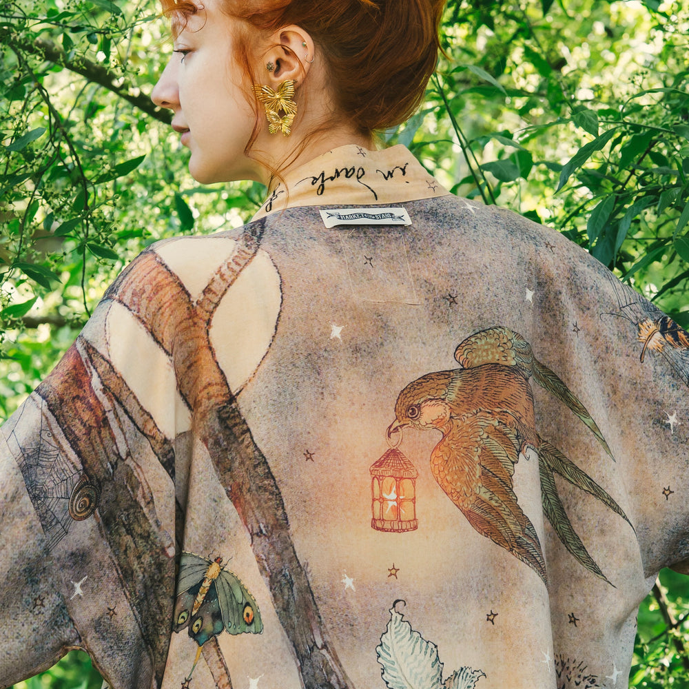 
                  
                    Model wearing an enchanting cropped kimono duster featuring illustrations by Charles van Sandwyck's, crafted from airy bamboo-based viscose with a touch of whimsical magic - Market of Stars
                  
                