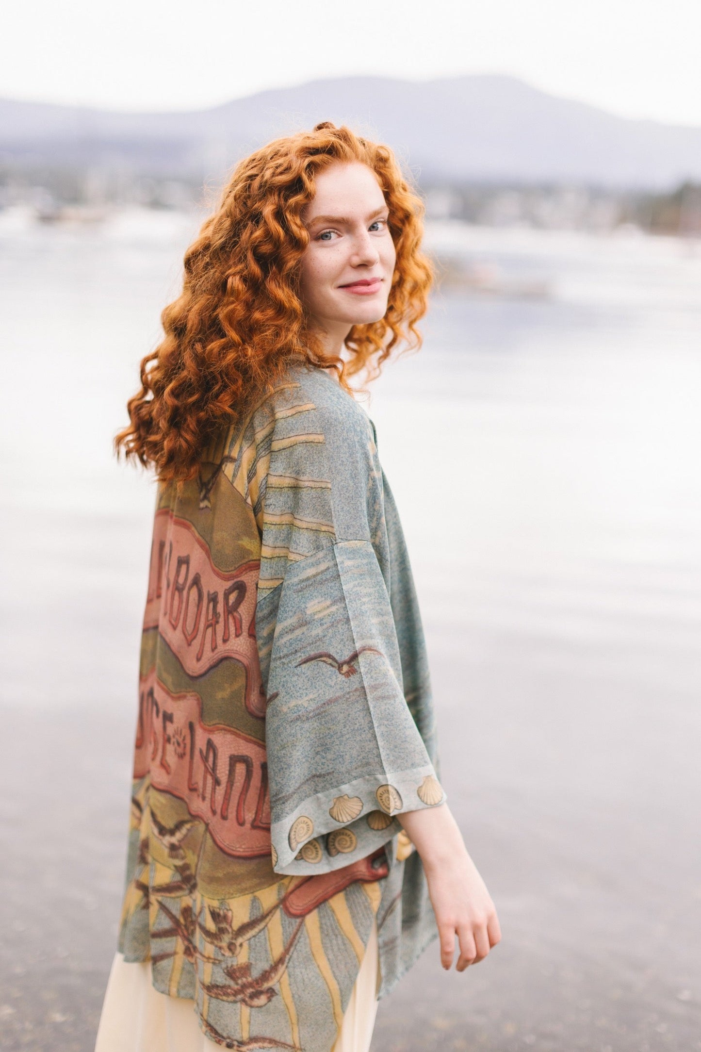 Lightweight duster featuring antique sunrise sky, mermaid, shell, and soaring birds artwork, shown on model at the beach – Market of Stars