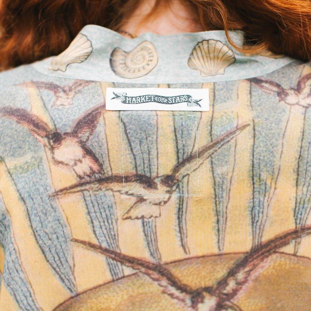 
                  
                    Lightweight duster featuring antique sunrise sky, mermaid, shell, and soaring birds artwork, shown on model at the beach – Market of Stars
                  
                